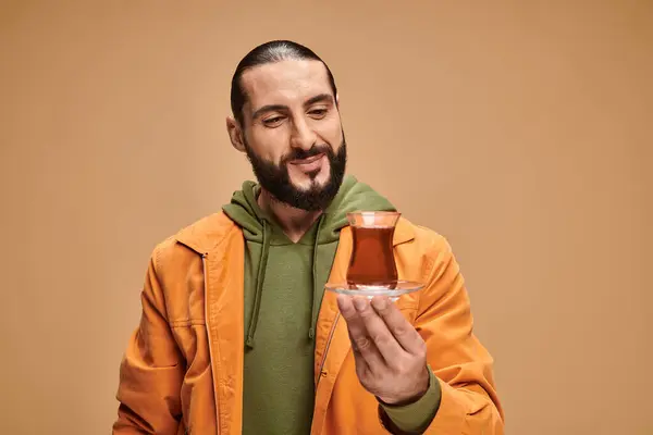 Felice uomo barbuto in abbigliamento casual guardando il tè turco in tazza di vetro su sfondo beige — Foto stock
