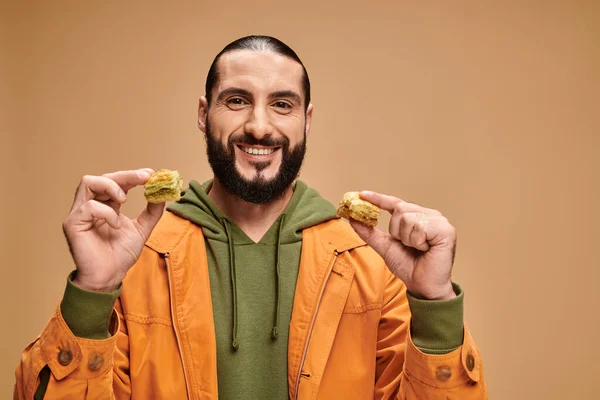 Hombre barbudo feliz sosteniendo dos tipos diferentes de baklava sobre fondo beige, delicias turcas - foto de stock