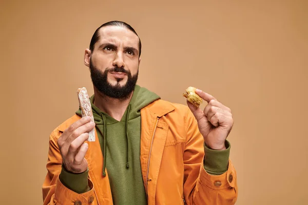 Hombre barbudo confundido sosteniendo baklava y cevizli sucuk sobre fondo beige, delicias turcas - foto de stock