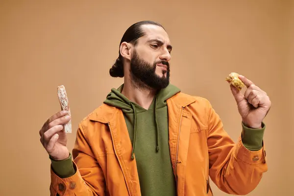 Confuso uomo barbuto arabo che tiene baklava e cevizli sucuk su sfondo beige, delizie turche — Foto stock