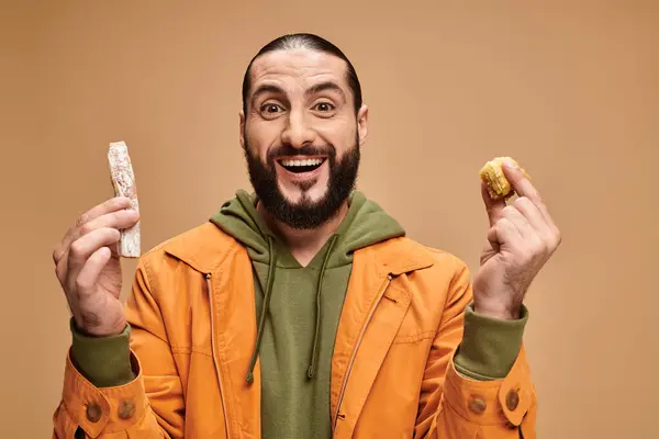 Excited arabic bearded man holding baklava and cevizli sucuk on beige backdrop, turkish delights — Stock Photo