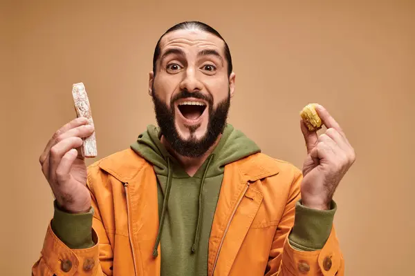 Excited arabic bearded man holding baklava and cevizli sucuk on beige backdrop, turkish delights — Stock Photo