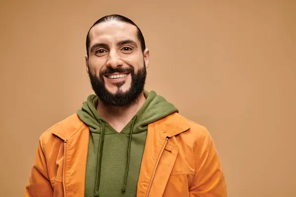 Hombre árabe feliz con barba de pie en traje casual y mirando a la cámara en el fondo beige - foto de stock