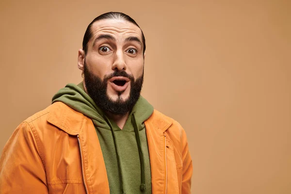 Überraschter arabischer Mann mit Bart steht in legerer Kleidung vor beigem Hintergrund und blickt in die Kamera — Stockfoto