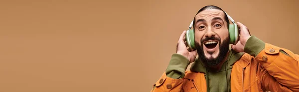 Cheerful middle eastern man with beard listening music in wireless headphones on beige, banner — Stock Photo