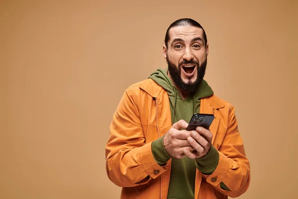 Glücklicher Mann aus dem Nahen Osten mit Bart, Smartphone auf beigem Hintergrund, digitales Zeitalter — Stockfoto