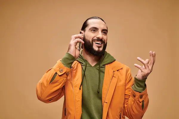 Счастливый ближневосточный мужчина с бородой разговаривает на смартфоне на бежевом фоне, цифровой век — стоковое фото