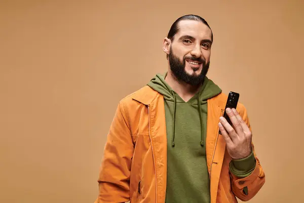 Щасливий східний чоловік з бородою тримає смартфон на бежевому фоні, цифровий вік — стокове фото