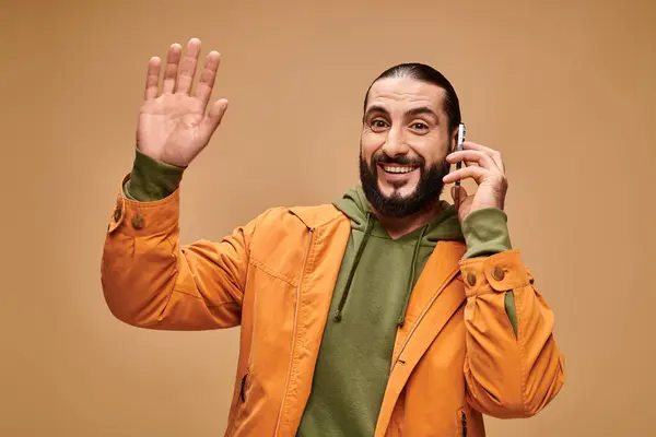 Amigável homem do Oriente Médio com barba falando no smartphone no fundo bege, mão onda — Fotografia de Stock