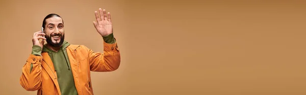 Friendly middle eastern man with beard talking on smartphone on beige background, wave hand banner — Stock Photo
