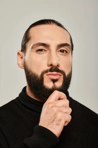Bellissimo uomo mediorientale in dolcevita nero toccare la barba su sfondo grigio, fiducia — Foto stock