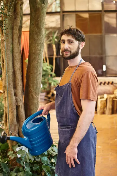 Good looking and bearded gardener in denim apron watering plants in greenhouse, horticulture concept — Stock Photo