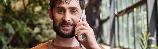 Hermoso jardinero barbudo en delantal sonriendo y hablando en el teléfono inteligente en invernadero, pancarta - foto de stock
