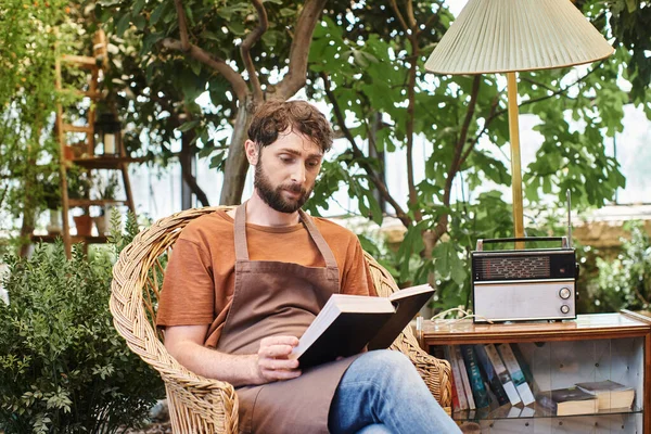Hermoso jardinero barbudo en delantal de lino lectura libro y sentado alrededor de las plantas en invernadero - foto de stock