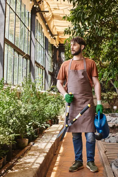 Jardineiro barbudo em luvas e avental segurando grande tesoura de jardinagem em torno de plantas em estufa — Fotografia de Stock