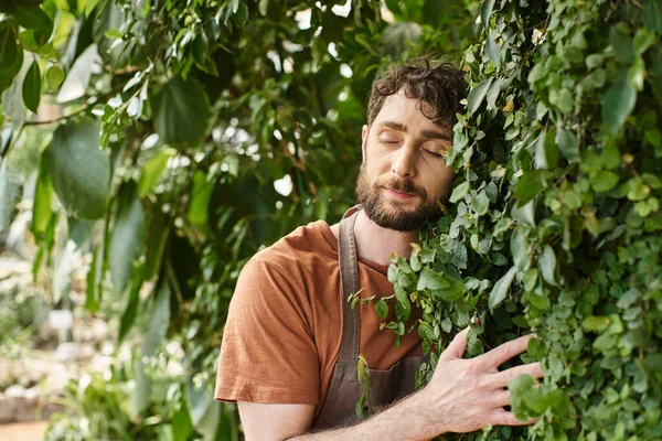 Jardineiro barbudo satisfeito em avental de linho sorrindo enquanto trabalhava em estufa, conceito de horticultura — Fotografia de Stock