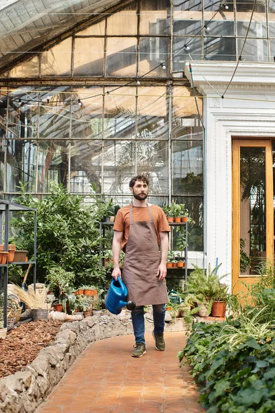 Jardineiro bonito e barbudo em avental segurando rega pode perto de plantas verdes em estufa — Fotografia de Stock