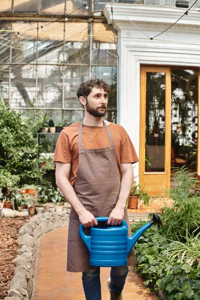 Красивый и бородатый садовник в фартуке держит поливальную банку рядом с зелеными растениями в теплице — стоковое фото