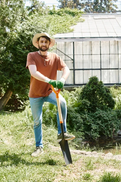 Cheerful farmer with beard wearing sun hat and standing with shovel near plants and greenhouse — Stock Photo