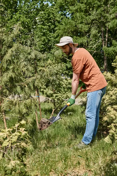 Giardiniere allegro con barba che indossa cappello da sole e scavo con pala vicino a piante e serra — Foto stock