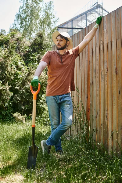 Handsome farmer with beard wearing sun hat and standing with shovel near plants and fence — Stock Photo