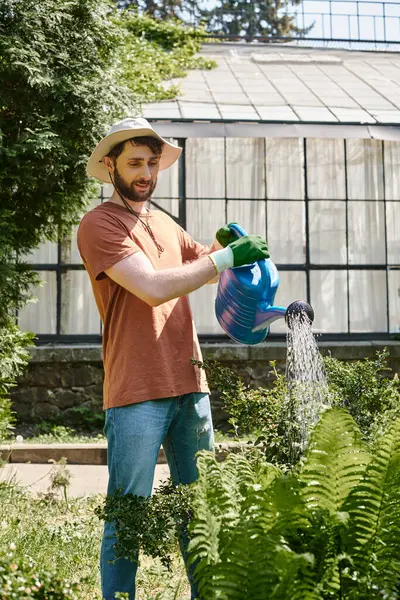 Hermoso y barbudo jardinero en sombrero de sol regar plantas cerca de invernadero en el campo - foto de stock