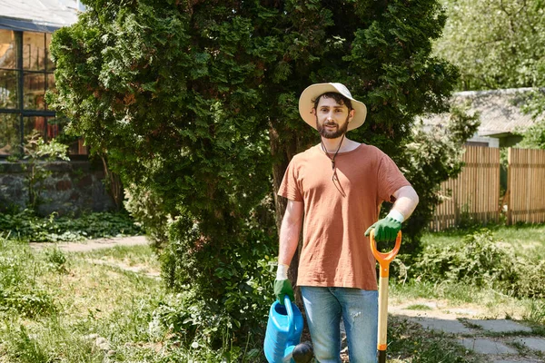 Handsome and bearded gardener in sun hat holding watering can and shovel in countryside — Stock Photo