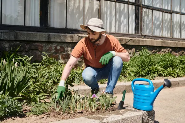 Handsome and bearded gardener in sun hat sitting near watering can and shovel in countryside — Stock Photo