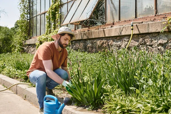 Handsome and bearded gardener in sun hat examining plant and sitting near watering can — Stock Photo