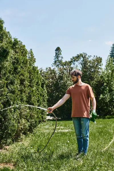 Bearded gardener in casual attire holding gloves and watering green lawn while working outdoors — Stock Photo