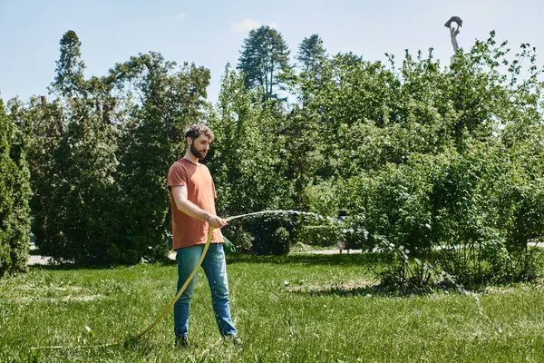 Бородатый садовник в повседневной одежде, держащий перчатки и поливающий зеленую лужайку во время работы на открытом воздухе — стоковое фото