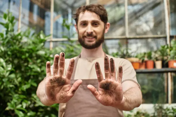 Heureux barbu jardinier dans tablier montrant ses mains sales après avoir travaillé avec des plantes et de la terre — Photo de stock