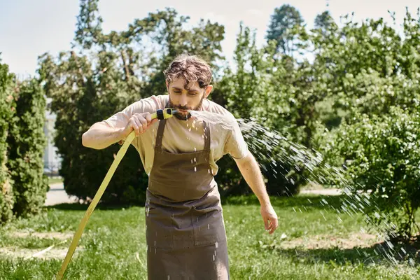 Bearded gardener in linen apron drinking water from hose after working in garden, candid photo — Stock Photo