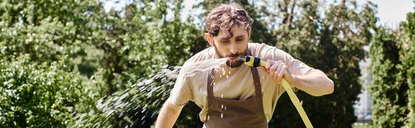 Jardinier barbu dans le tablier de lin eau potable du tuyau après avoir travaillé dans le jardin, bannière franche — Photo de stock