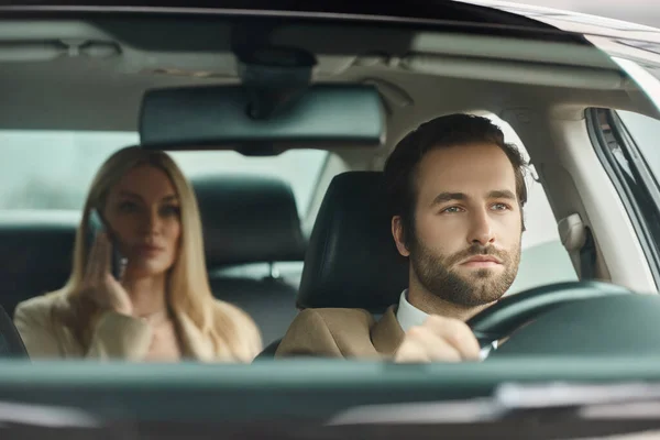 Concentrated bearded man driving luxury car with blonde businesswoman talking on mobile phone — Stock Photo