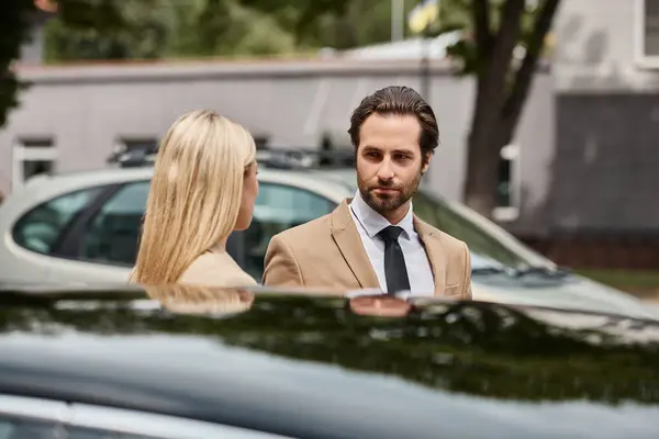 Guapo elegante hombre de negocios y mujer rubia mirándose el uno al otro cerca del coche en la calle urbana - foto de stock