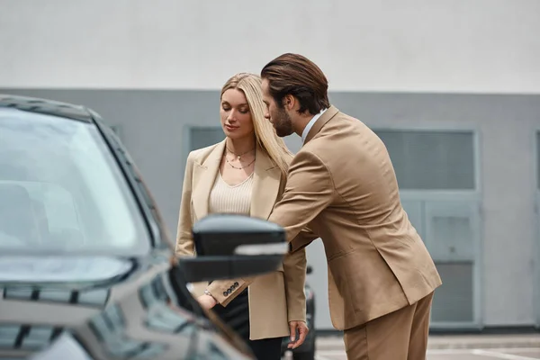 Elegante uomo barbuto apertura porta posteriore di auto di lusso vicino elegante donna d'affari bionda sulla strada — Foto stock