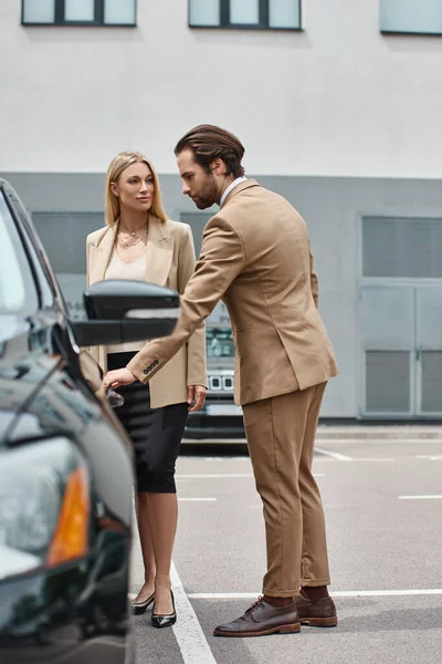 Hombre elegante guapo abriendo la puerta trasera del coche de lujo cerca de la elegante mujer de negocios rubia en la calle - foto de stock