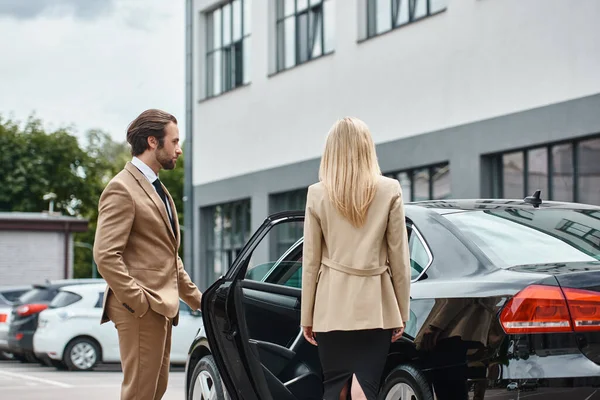 Elegante hombre de traje abriendo la puerta trasera del coche de lujo cerca de rubia exitosa mujer de negocios en la calle - foto de stock