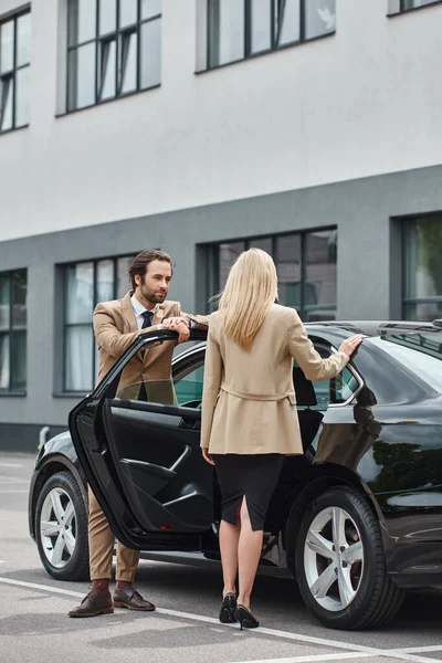 Uomo elegante in tuta apertura porta posteriore di auto di lusso vicino bionda donna d'affari di successo sulla strada — Foto stock