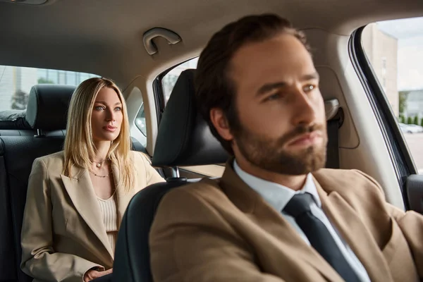 Desenfocado elegante hombre en formal desgaste conducir coche de lujo con elegante mujer de negocios rubia en asiento trasero - foto de stock