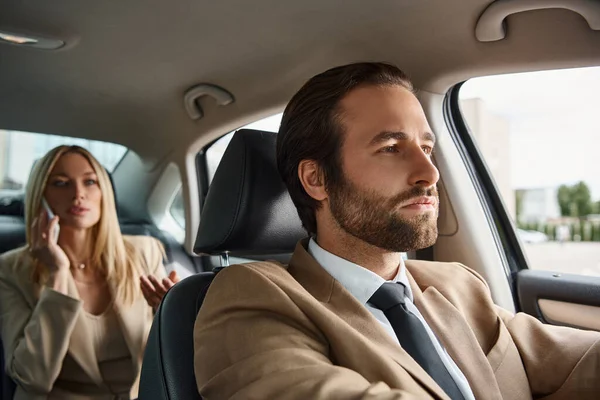 Hombre barbudo en traje de conducción de coches de lujo con elegante mujer de negocios rubia hablando en el teléfono inteligente - foto de stock