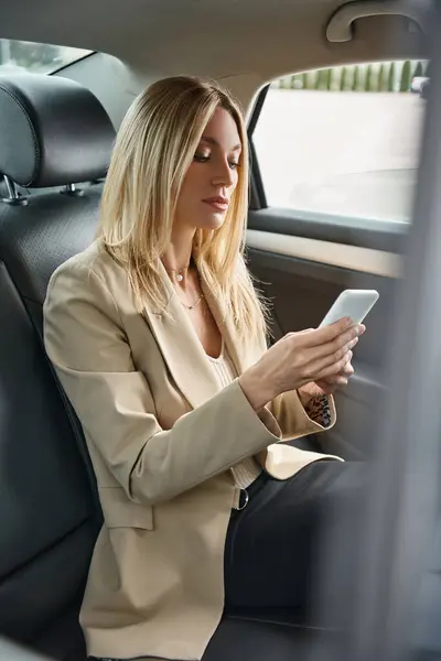 Stylish blonde businesswoman in formal wear messaging on mobile phone in luxury car on city street — Stock Photo