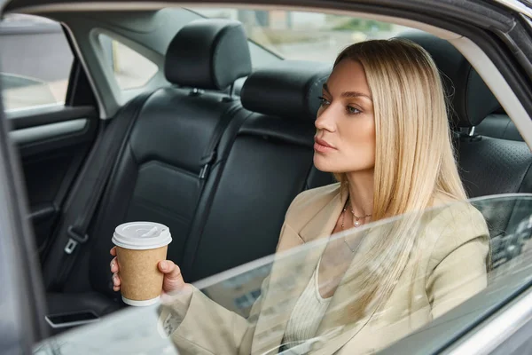 Attrayant blonde femme d'affaires en tenue formelle assis avec du café pour aller en voiture de luxe sur la rue — Photo de stock