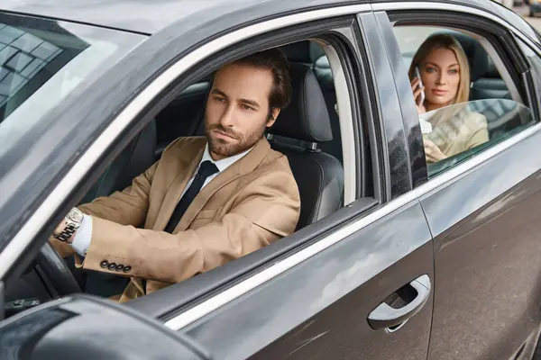 Hombre guapo y elegante coche de conducción cerca de atractiva mujer de negocios rubia hablando en el teléfono inteligente - foto de stock