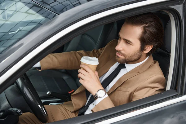 Confiado hombre de negocios barbudo en traje elegante beige sosteniendo café para ir y conducir un coche en la calle - foto de stock