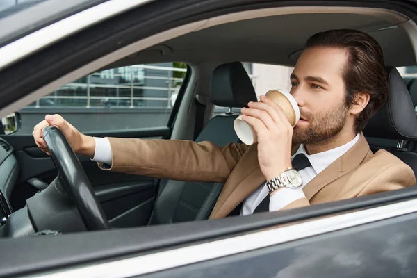 Красивый бородатый бизнесмен в бежевом элегантном костюме пьет кофе, чтобы поехать и водить машину по улице — стоковое фото