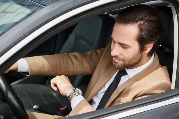 Красивый бородатый бизнесмен в формальной одежде смотрит на наручные часы во время вождения автомобиля на улице — стоковое фото