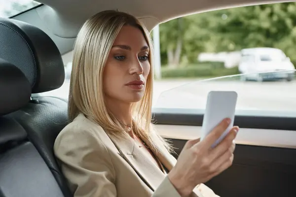 Mujer de negocios rubia en elegante desgaste formal navegar por Internet en el teléfono móvil mientras está sentado en el coche - foto de stock