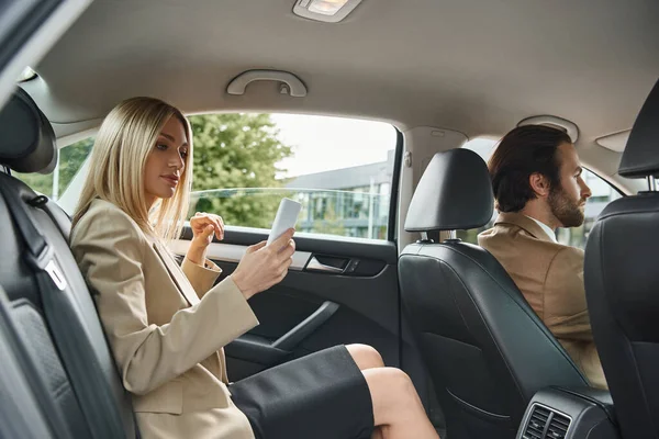 Blonde élégante femme d'affaires en utilisant smartphone près de l'homme élégant conduisant voiture de luxe sur la rue — Photo de stock